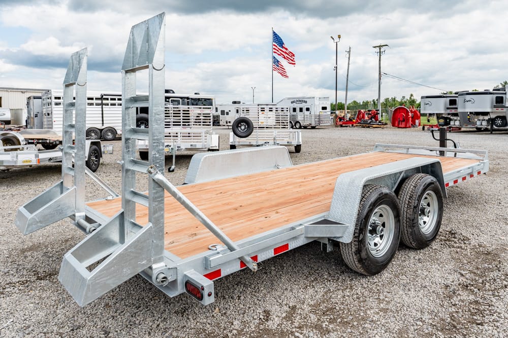 Double-axle utility trailer with a wood bed and fold-down ramps in a gravel lot.