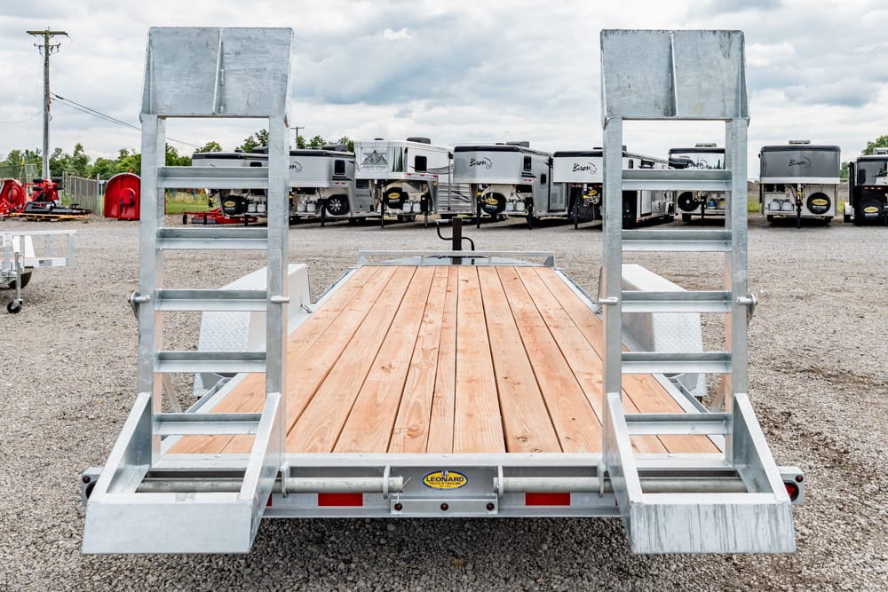 Back view of a flatbed trailer with wooden deck and dual metal ramps, parked on gravel with RVs in the background.