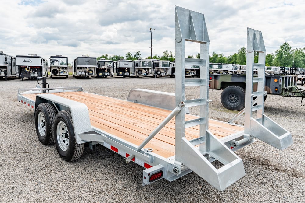 Flatbed trailer with wooden deck and raised dual ramps in a gravel lot of trailers behind it