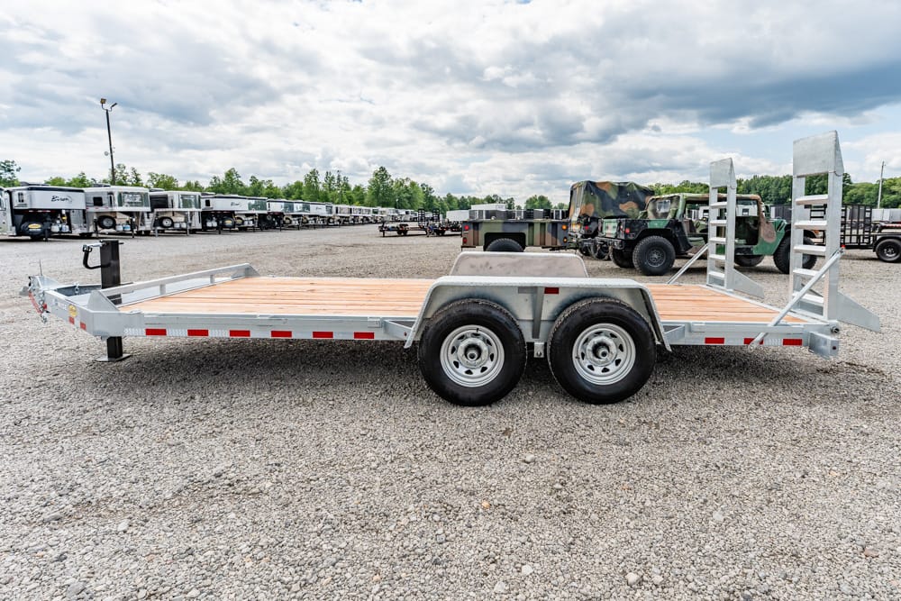 Dual-axle flatbed trailer with wooden deck on a gravel lot, featuring folded rear ramps and a front hitch area. The trailer is parked among a line of other trailers and military vehicles in the background.