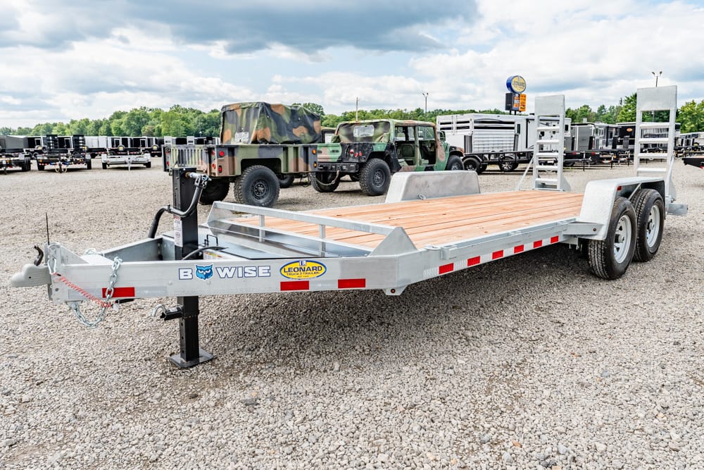 Long aluminum flatbed trailer with a wooden deck and dual axles parked on a gravel lot, with various trailers and trucks in the background.
