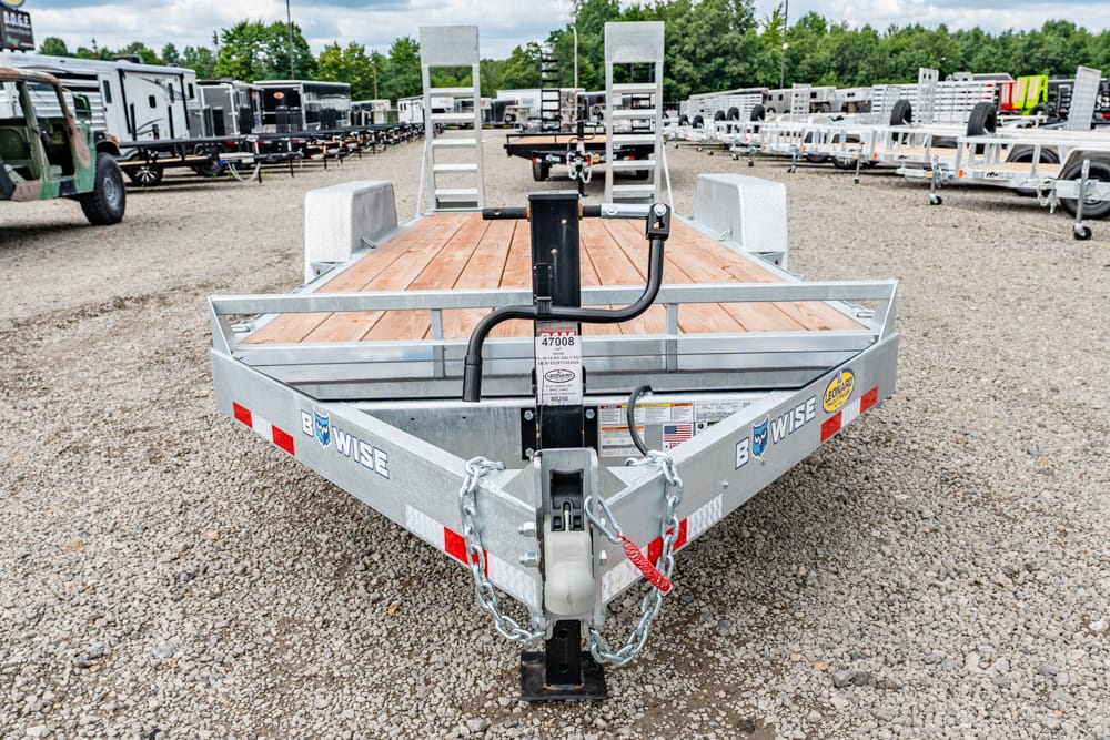 Flatbed trailer with a wooden deck and metal rails in a gravel lot; hitch and safety chains visible, with other trailers in the background.