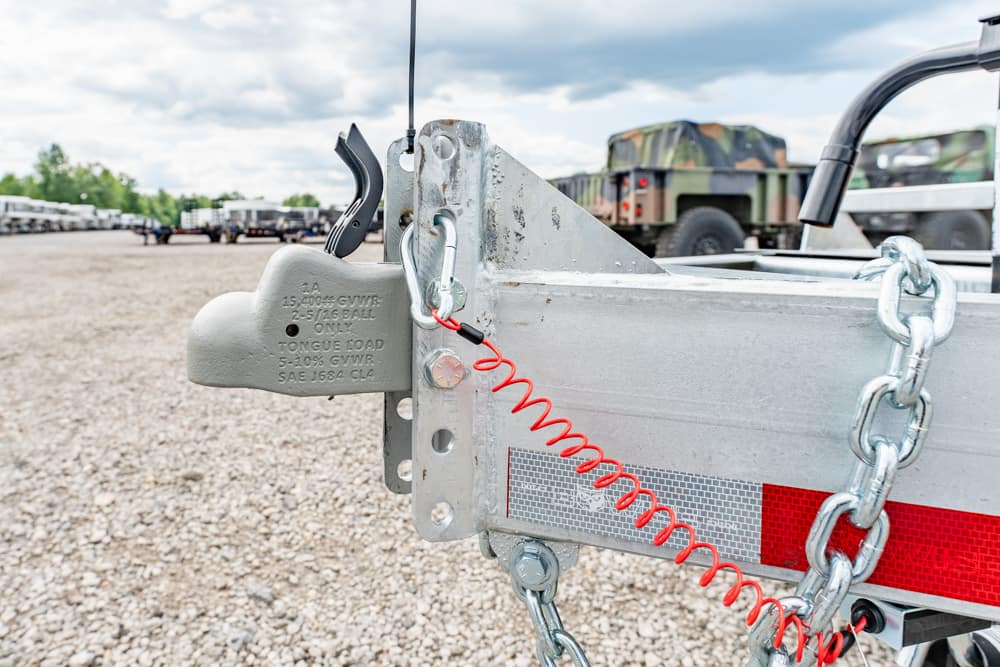 Close-up of a trailer hitch with safety chains and a red coiled tether on a gravel lot, with military trucks in the background.