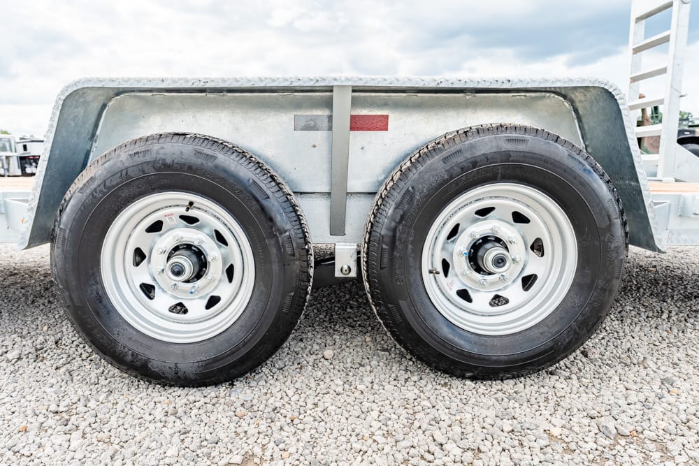 Dual wheels of a utility trailer on gravel, with metal fenders and frame visible in the scene.