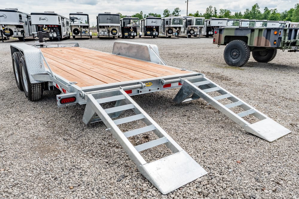 Flatbed trailer with two metal ramps extended, parked on gravel lot with other trailers in background