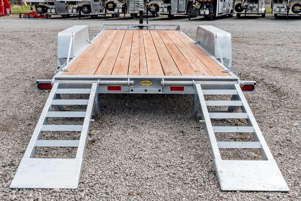 Flatbed trailer with a wooden deck and two metal loading ramps folded down on a gravel lot.