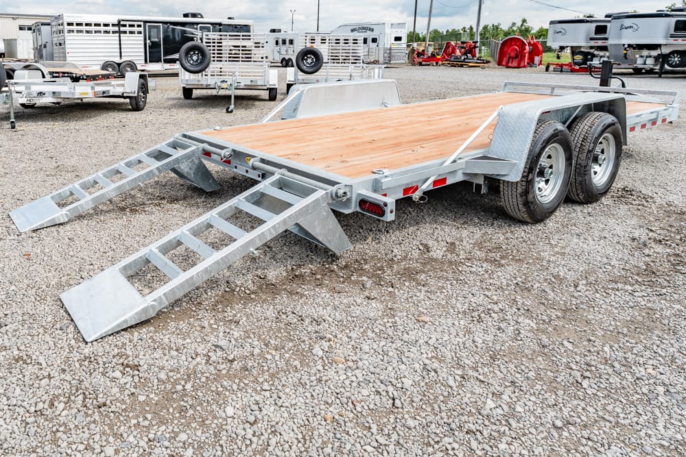 Dual-axle flatbed trailer with wood deck and two loading ramps extended on a gravel lot; other trailers in background.