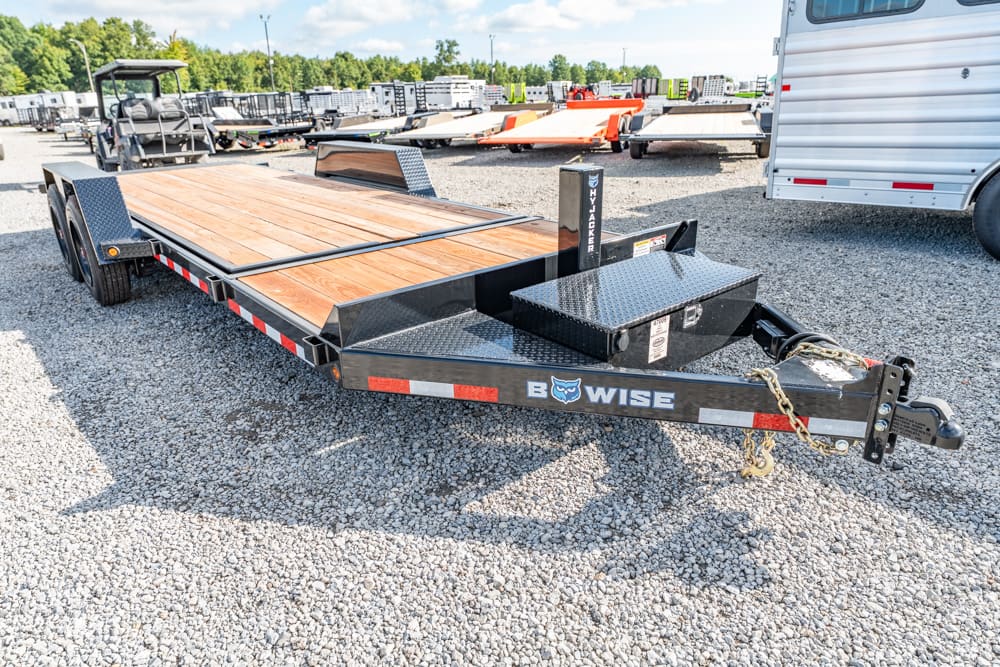 Black BEWISE flatbed trailer with a wooden deck parked in a gravel lot, with other trailers and a horse trailer in the background.