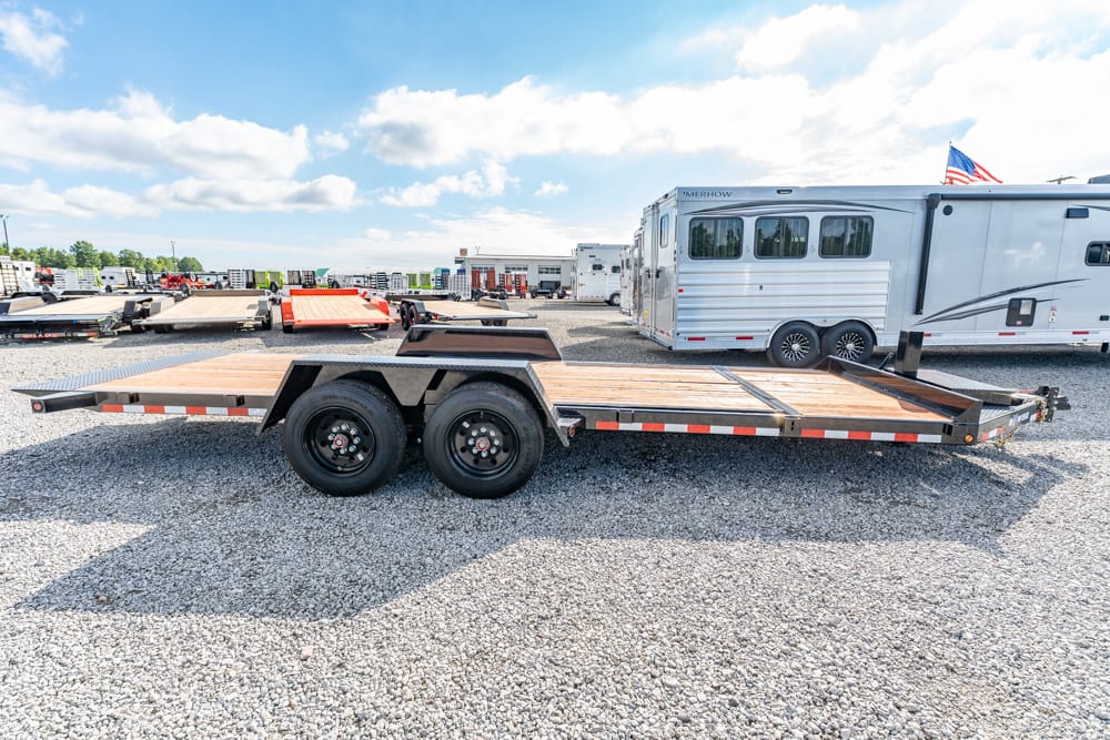 Dual-axle flatbed trailer on a gravel lot with an RV trailer in the background; outdoor trailer yard scene.