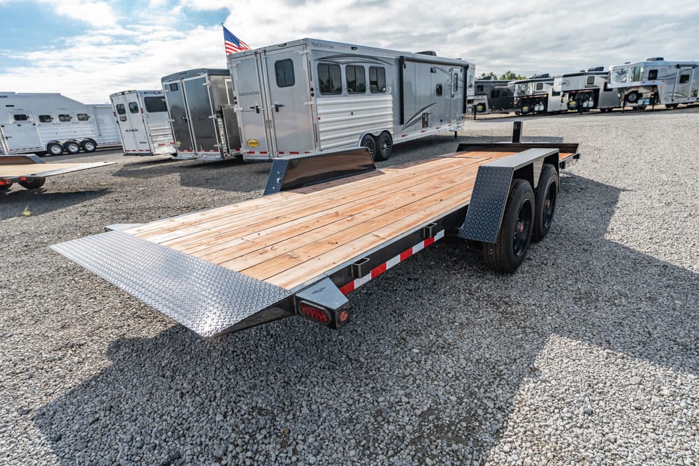 Flatbed trailer with wooden deck and dual-axle wheels parked in a gravel lot among RVs and trailers