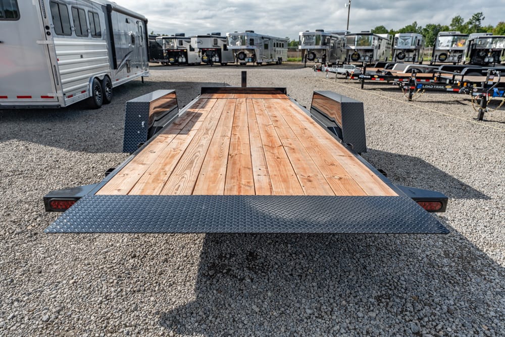 Flatbed trailer with a wooden deck and diamond-plate metal fenders parked on a gravel lot, with other trailers in the background.