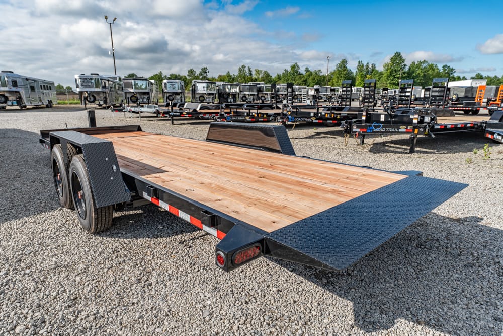 Dual-axle flatbed trailer with a wooden deck and a folding ramp, parked on a gravel lot with other trailers in the background.