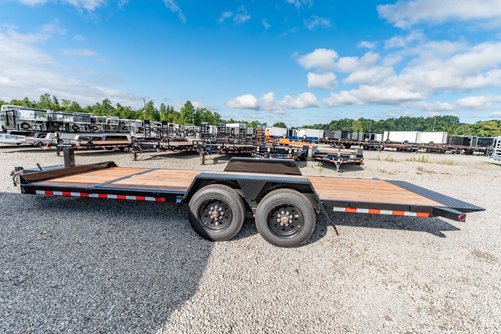Black flatbed trailer with wooden deck and dual axles in a gravel lot, with other trailers visible in the background under a blue sky.
