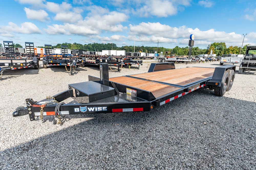 Black gooseneck flatbed trailer with a wooden deck, parked on a gravel lot among other trailers, under a blue sky with clouds.