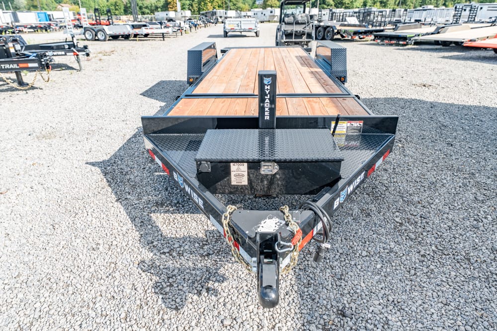 Flatbed utility trailer with wooden deck and black metal frame in a trailer lot, front hitch visible.
