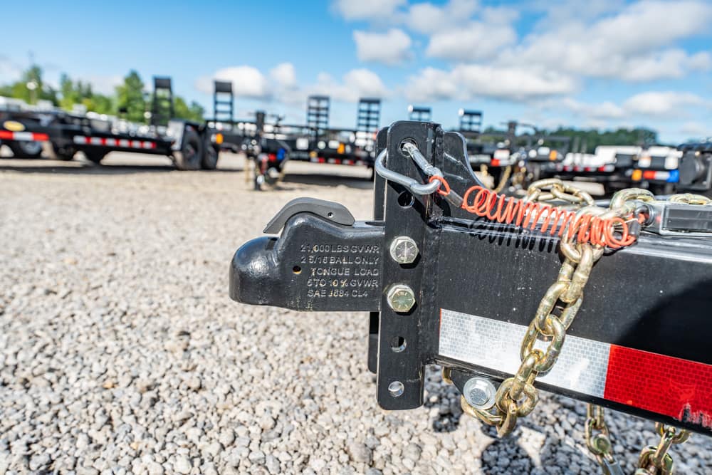 Close-up of a trailer hitch with safety chains and an orange coiled cable on a gravel lot, trucks in the background.