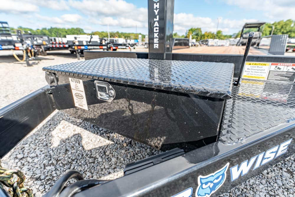 Black trailer with a diamond-plate toolbox mounted on the bed, warning labels visible, on a gravel lot with other trailers in the background.