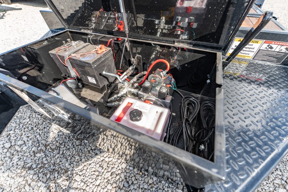 Open compartment of a trailer showing a battery, hydraulic pump, and wiring on a gravel surface.