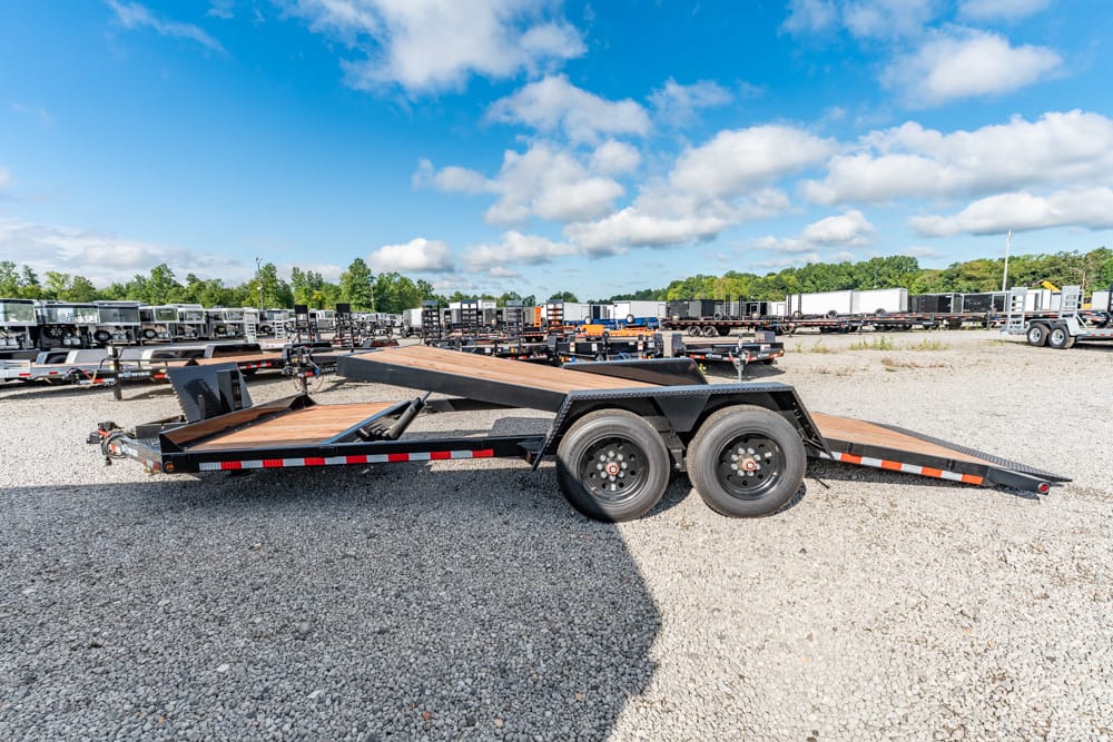 Dual-axle car hauler trailer with a raised ramp, parked on a gravel lot with many trailers nearby under a blue sky.