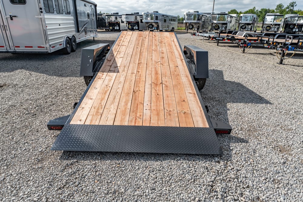 Flatbed trailer with a wooden deck and diamond-plate front ramp, parked on a gravel lot among other trailers.