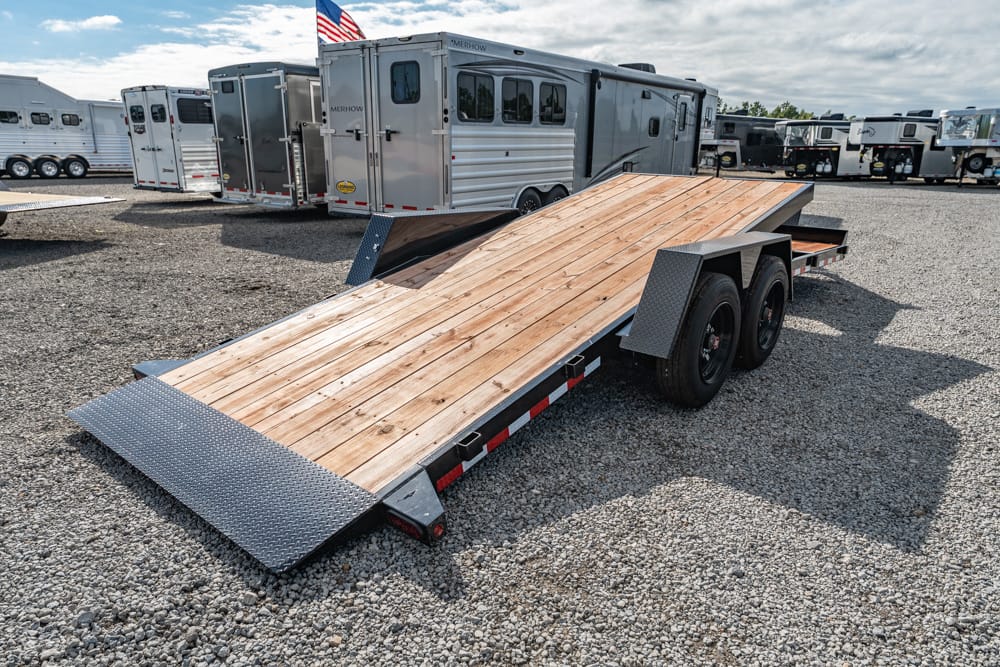 Two-axle utility trailer with a long wooden ramp resting on a gravel lot, background filled with horse trailers and rigs.