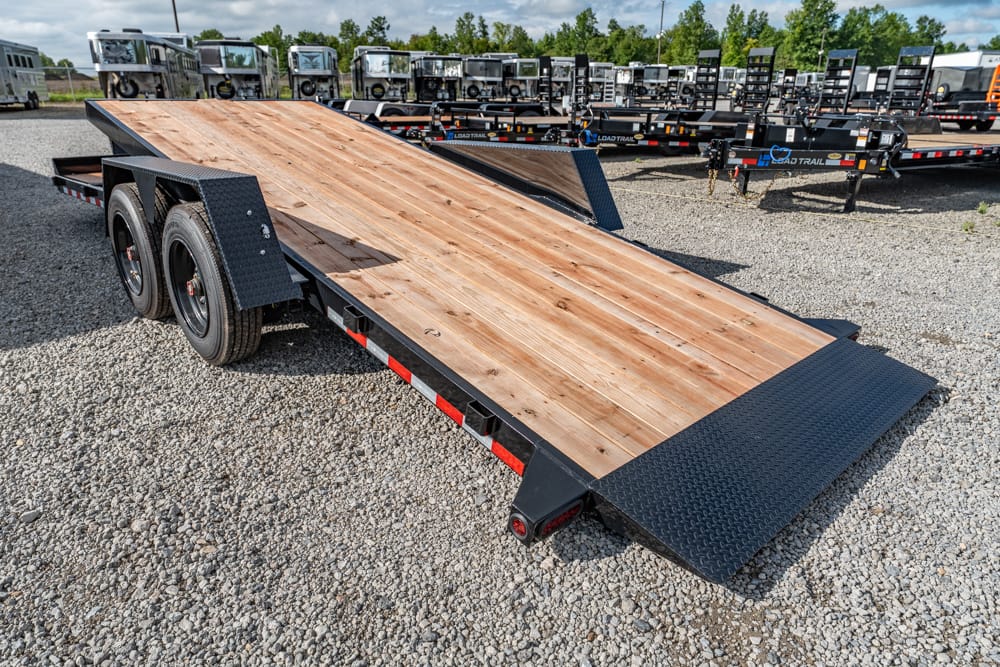 Black tandem-axle car trailer with a wooden deck and folding ramps on a gravel lot, ready for hauling vehicles/surfaced with red-and-white reflective stripes.