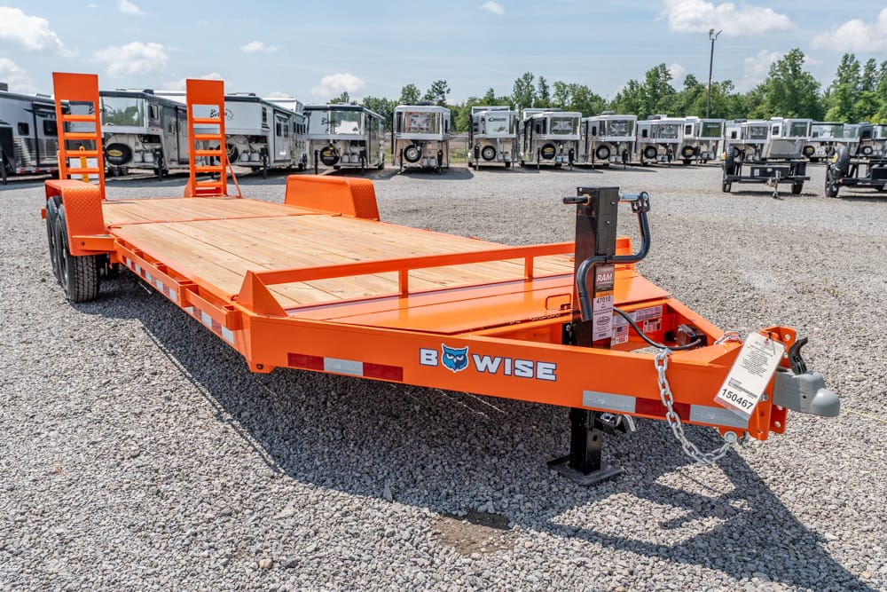 Orange B-Wise equipment trailer with wooden deck and rear ramps on a gravel lot, backed by a row of similar trailers and equipment in the background.