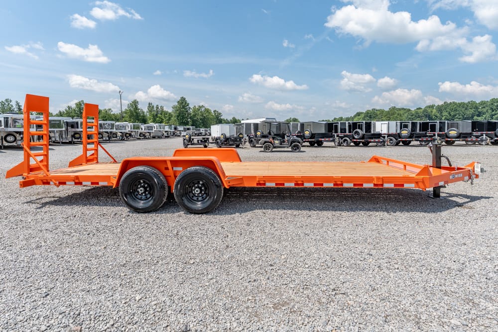 Orange car hauler trailer with dual axles and rear ramps on a gravel lot, surrounded by other trailers under a blue sky.