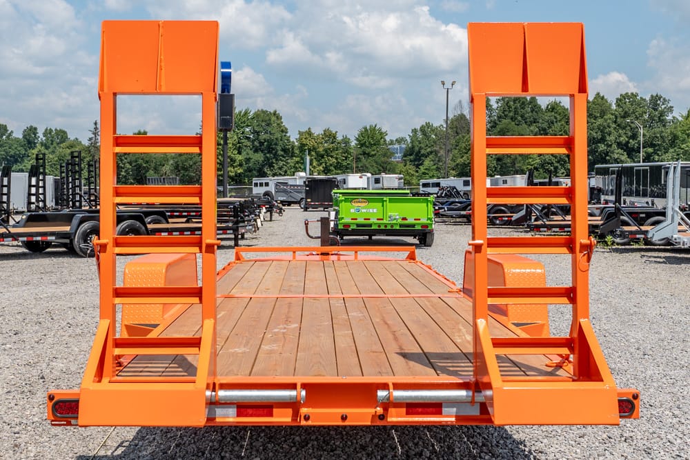 Bright orange equipment trailer with tall ramps and a wooden deck in a gravel trailer yard, with other trailers in the background at distance.