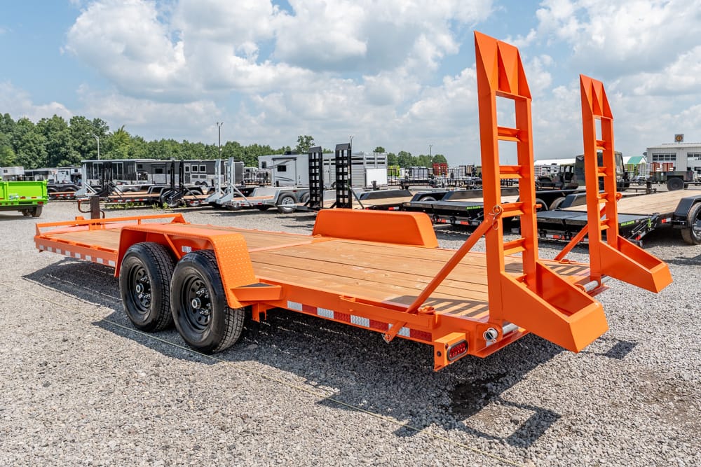 Orange flatbed trailer with two tall rear loading ramps raised, parked on a gravel lot with other trailers nearby.