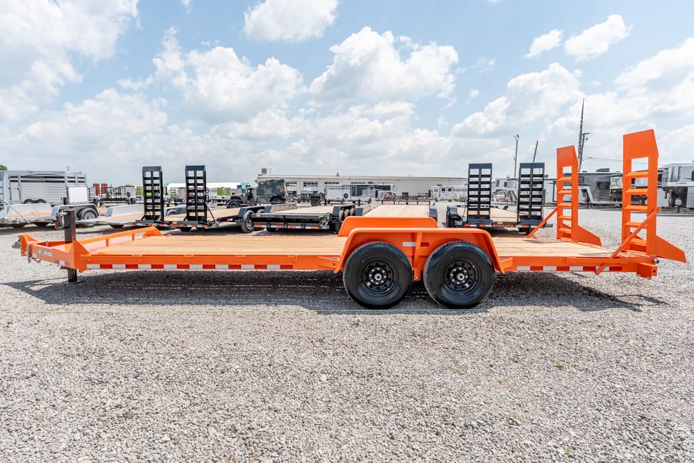 Orange flatbed equipment trailer with dual axles and tall rear ramps parked on a gravel lot.