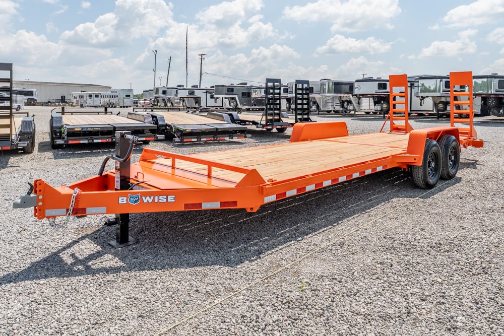Orange flatbed equipment trailer with dual rear ramps and two axles on a gravel lot, with other trailers in the background (brand: B-WISE).