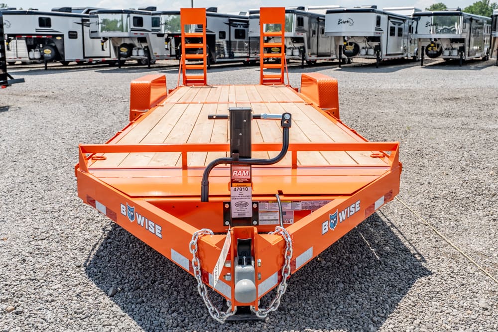 Orange equipment trailer with wooden deck and safety chains, parked on a gravel lot with travel trailers in the background.