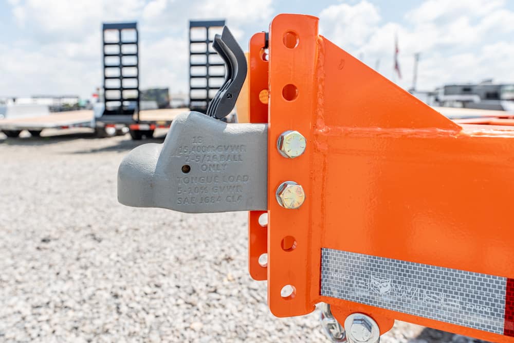 Close-up of a gray trailer coupler lock on an orange hitch, with bolts and reflective tape in a gravel lot.