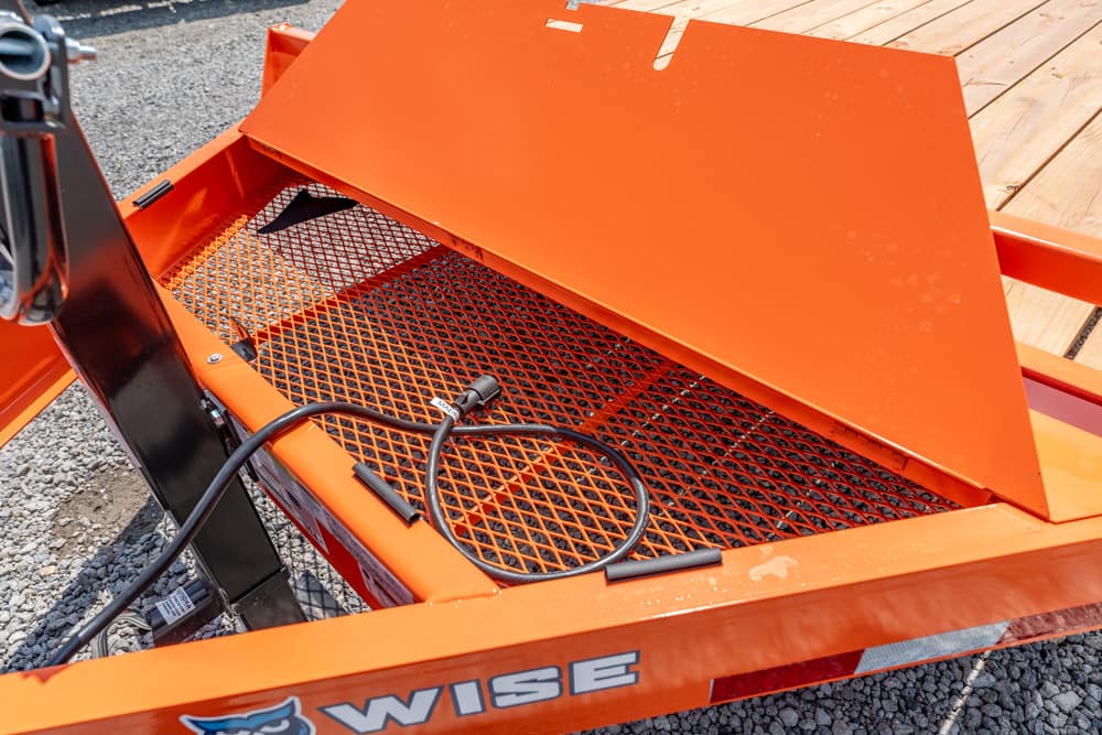 Orange utility trailer with a metal mesh floor, a coiled black power cable, and a folded ramp on a gravel surface.