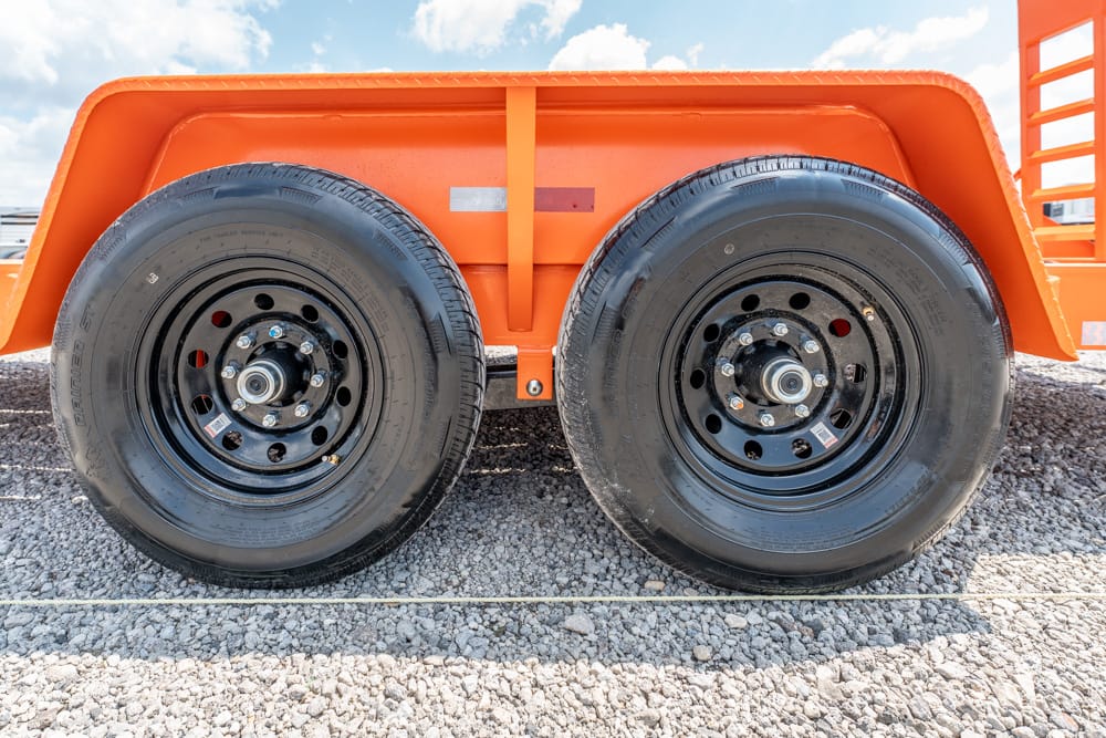Close-up of two black wheel rims with lug nuts on an orange trailer, resting on gravel under a blue sky with clouds.