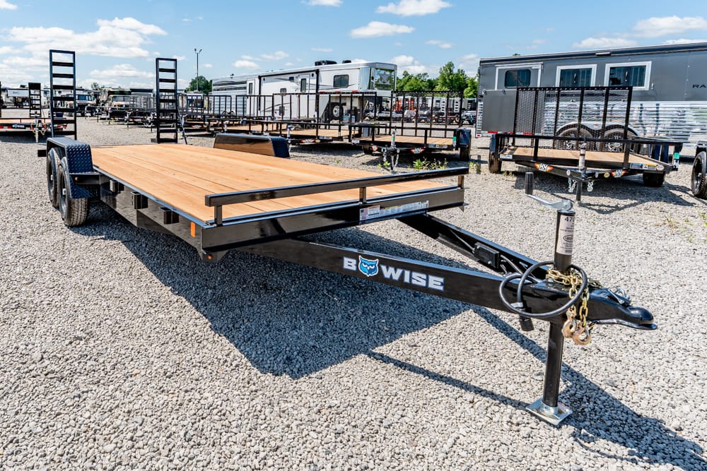 Flatbed trailer with wooden deck and black frame in a gravel trailer lot; hitch and chains visible in foreground, other trailers in background