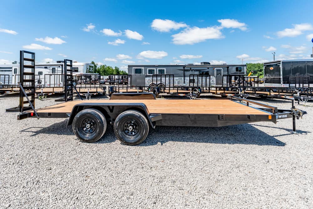 Flatbed trailer with a wooden deck and dual rear wheels parked on gravel lot, with other trailers and RVs in the background.