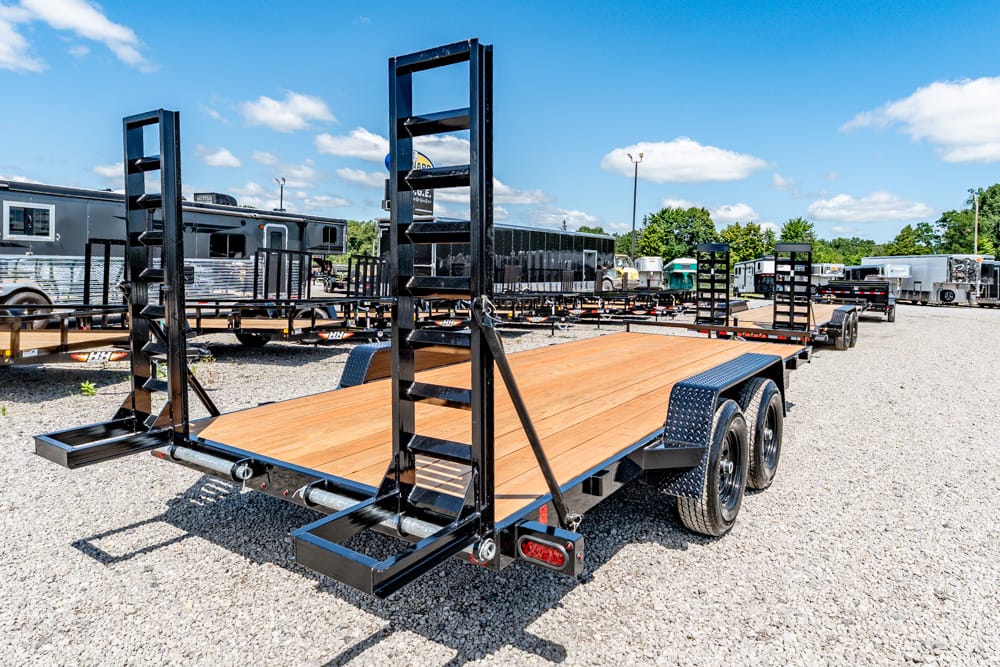 Black steel car hauler trailer with a wooden deck, dual axles, and tall ramp frames, parked on a gravel lot with other trailers in the background.