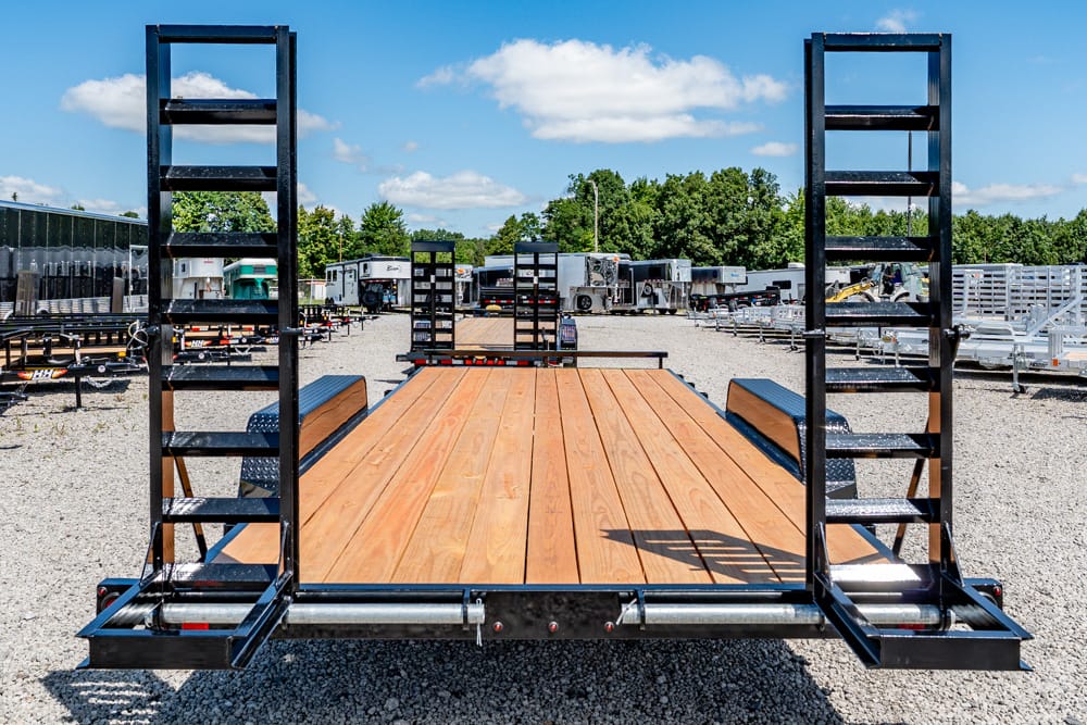 Flatbed trailer with a wooden deck and dual fold-down ramps, parked in an outdoor trailer lot under a blue sky.