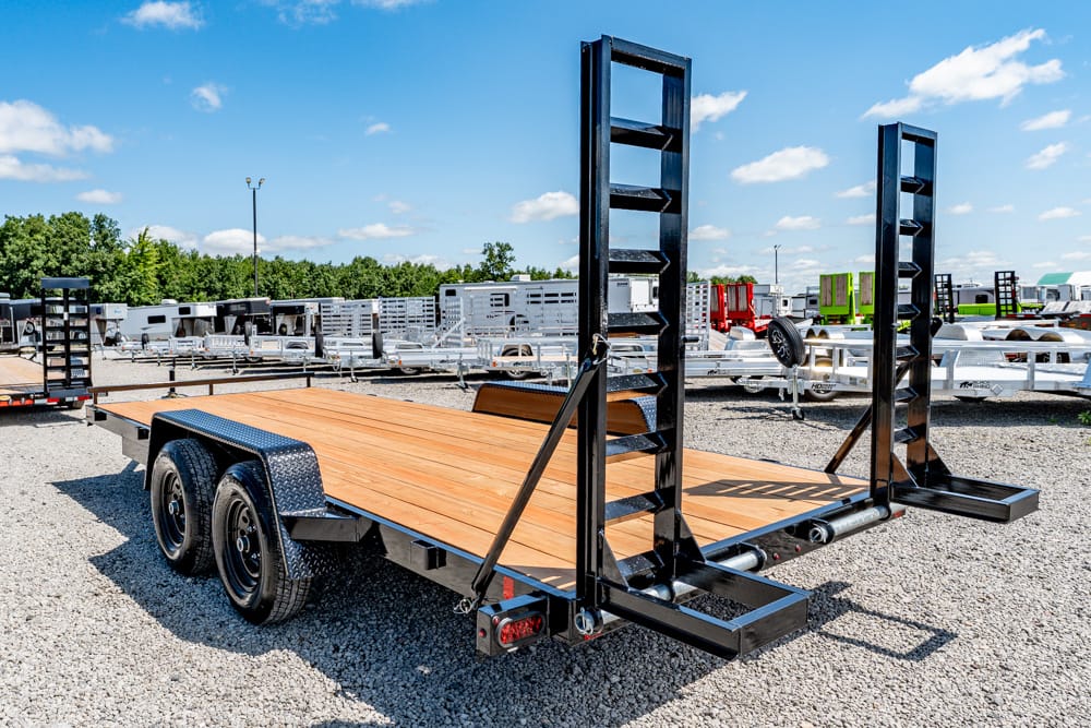 Flatbed trailer with wooden deck and tall loading ramps in a gravel lot, other trailers in background.