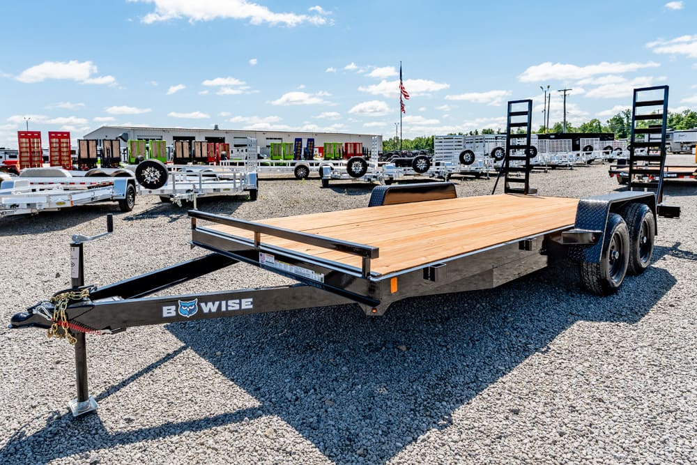 Flatbed equipment trailer with a wooden deck in a gravel lot, branded Bow-Wise, dual rear axles and ramps at the back used for hauling vehicles.