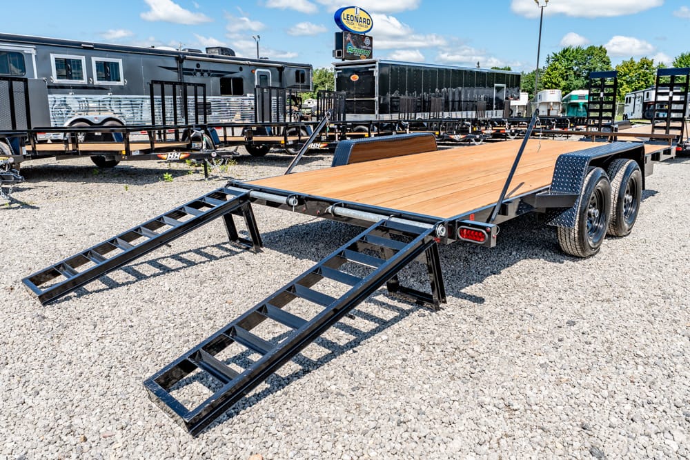Dual-axle flatbed trailer with a wood deck and two lowered metal ramps on a gravel lot, ready for loading.