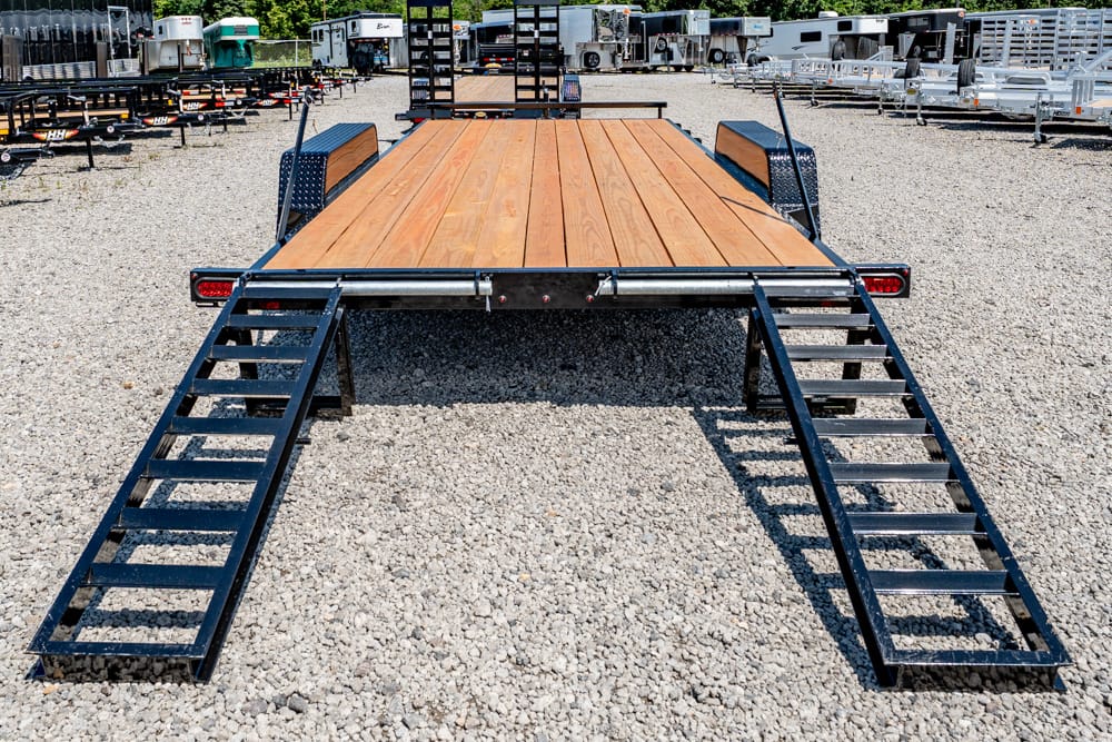 Flatbed trailer with a wooden deck and two retractable ramps extended for loading on a gravel lot.