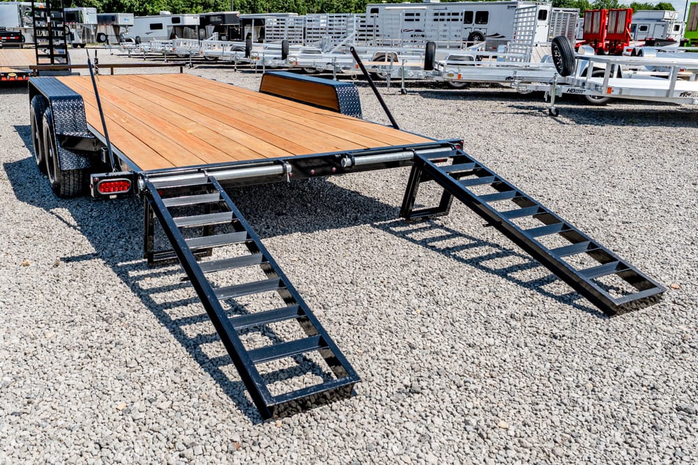 Dual-axle flatbed trailer with a wooden deck and two folding loading ramps extended on a gravel lot. Background shows more trailers and equipment.