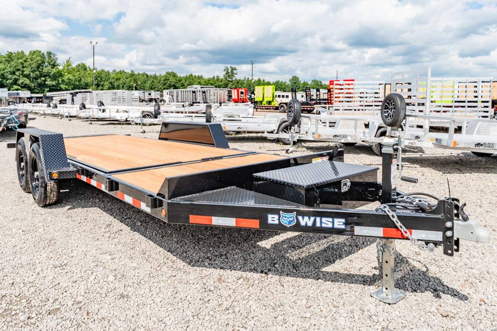 Flatbed trailer with wooden deck and black frame in a trailer yard; displays 'B-WISE' branding at the hitch area.
