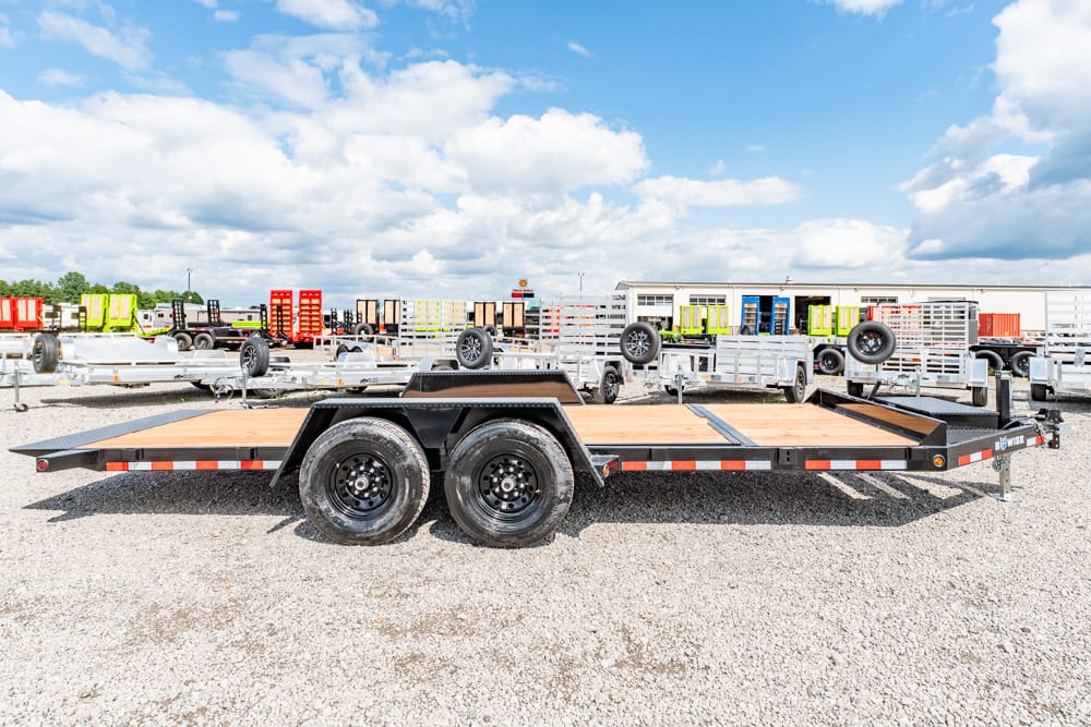 Flatbed trailer with dual rear wheels on a gravel lot, surrounded by other trailers and equipment on a sunny day.