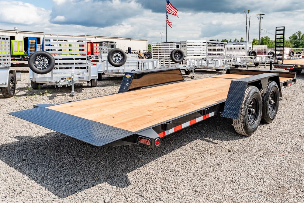 Dual-axle flatbed trailer with a wooden deck and diamond-plate ramps, parked on a gravel lot with other trailers and an American flag in the background.