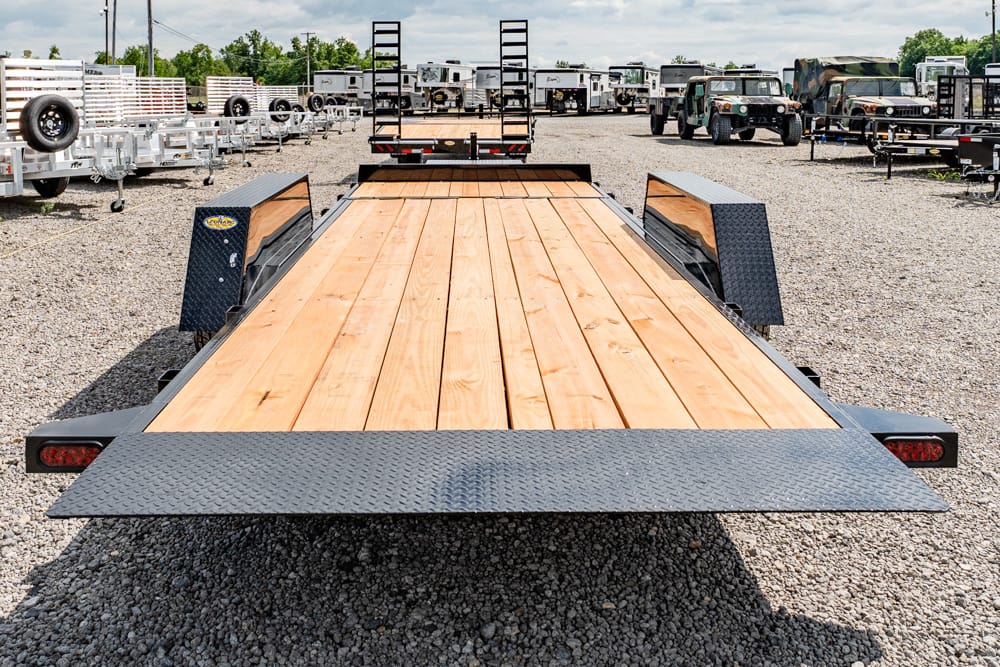 Flatbed trailer with a wooden deck and diamond-plate ramps parked on a gravel lot among other trailers and trucks