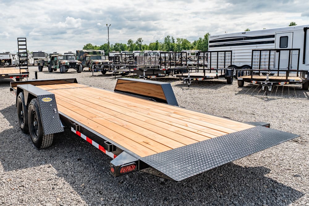 Flatbed car hauler trailer with a wooden deck and diamond-plate ramps in a gravel trailer lot